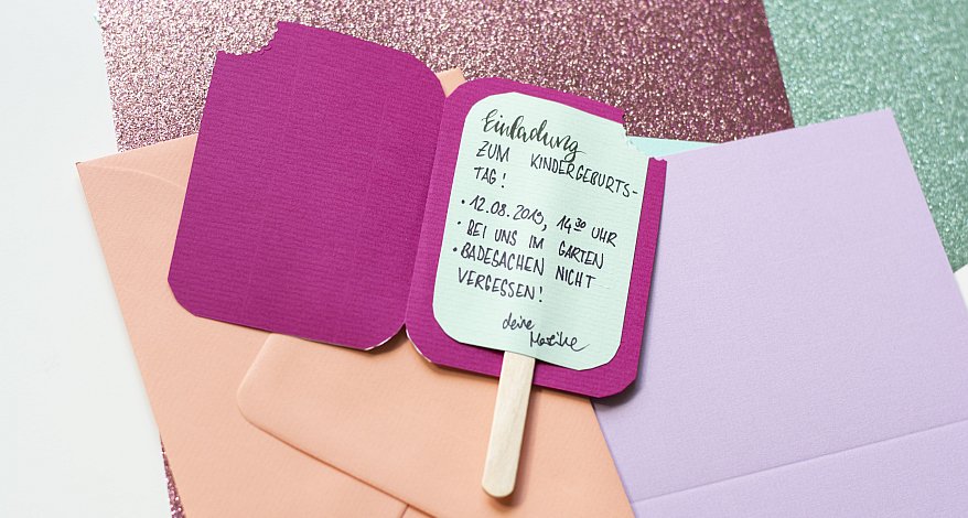 Eine Einladungskarte in Form eines Eis am Stiel liegt aufgeklappt auf verschiedenen Papieren von Rössler.