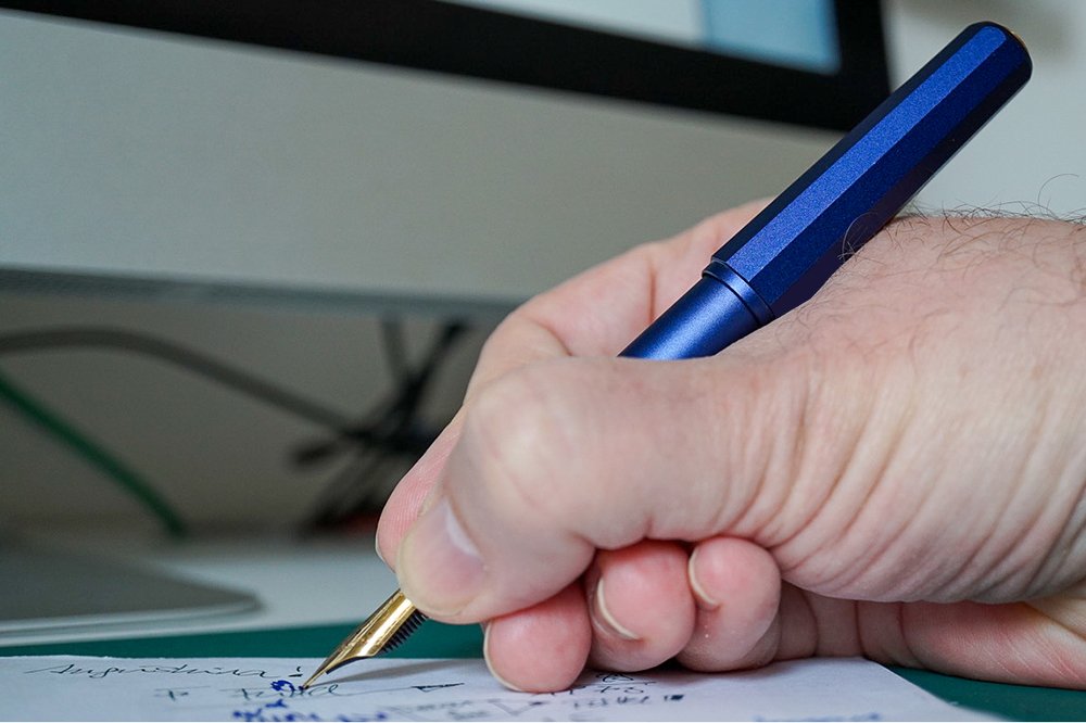 Eine Hand hält einen blauen Füller aus MEtall mit goldfarbener Spitze. Die Person mal mit dem Füller mit blauer Tinte etwas auf ein Blatt. Im Hintergrund ist unscharf ein Computerbildschirm erkennbar.