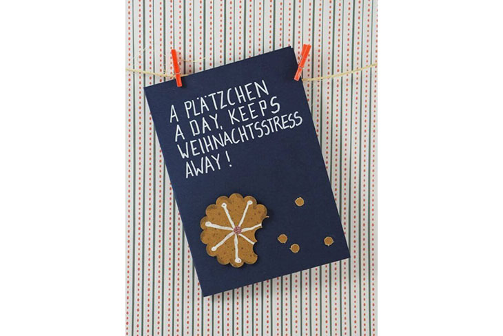 Eine blaue Karte, die den Text "A Plätzchen a day, keeps the Weihnachtsstress away!"zeigt, ist mit roten Holzklammern an einer Schnur befestigt. Darauf ist ein brauner Motivkarton zu sehen, der wie ein Plätzchen geformt und bemalt ist. Kleine braune Punkte sind als Kekskrümel am unteren Bildrand verteilt.
