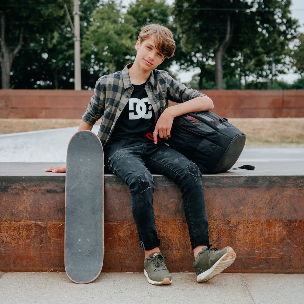 Ein Junge ist auf dem Skateplatz. Unter dem Arm hat er einen Rucksack der Marke satch. Im Vordergrund ist ein Skateboard zu sehen.