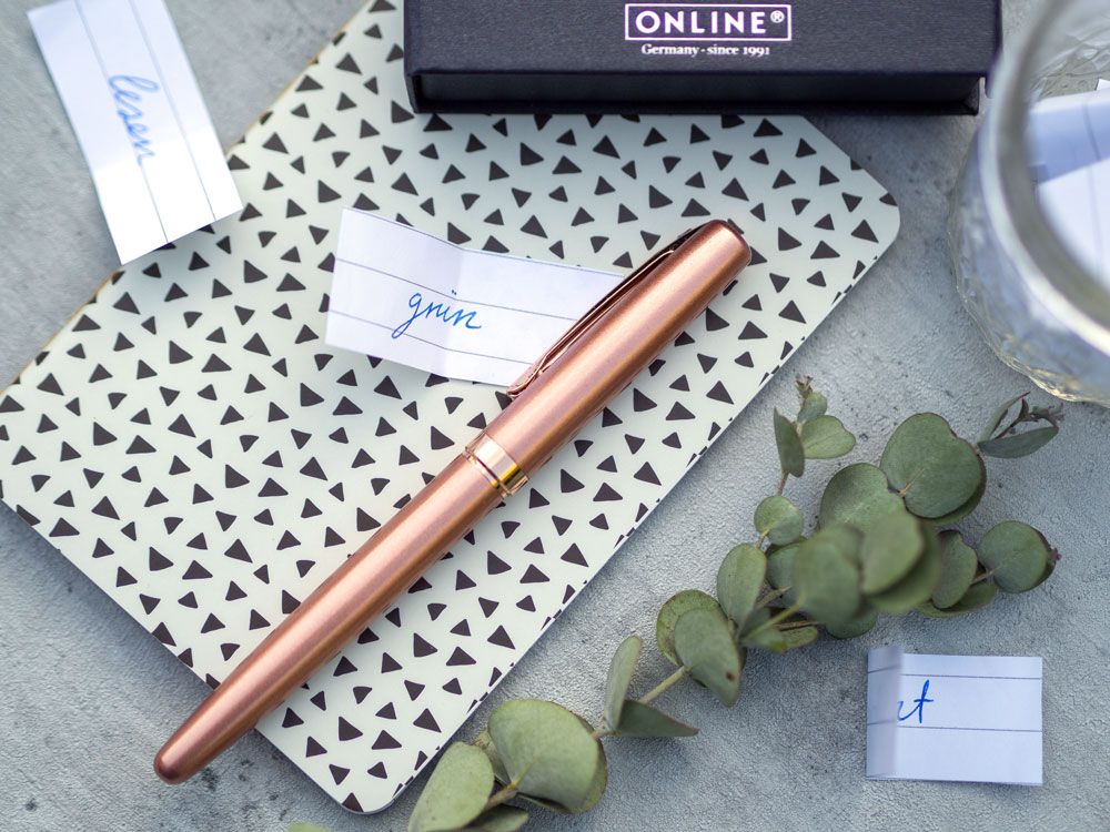 Rosegoldener Füllhalter der Marke Online auf Notizbuch mit Zetteln, Eukalyptuszweig und Schreibbox.