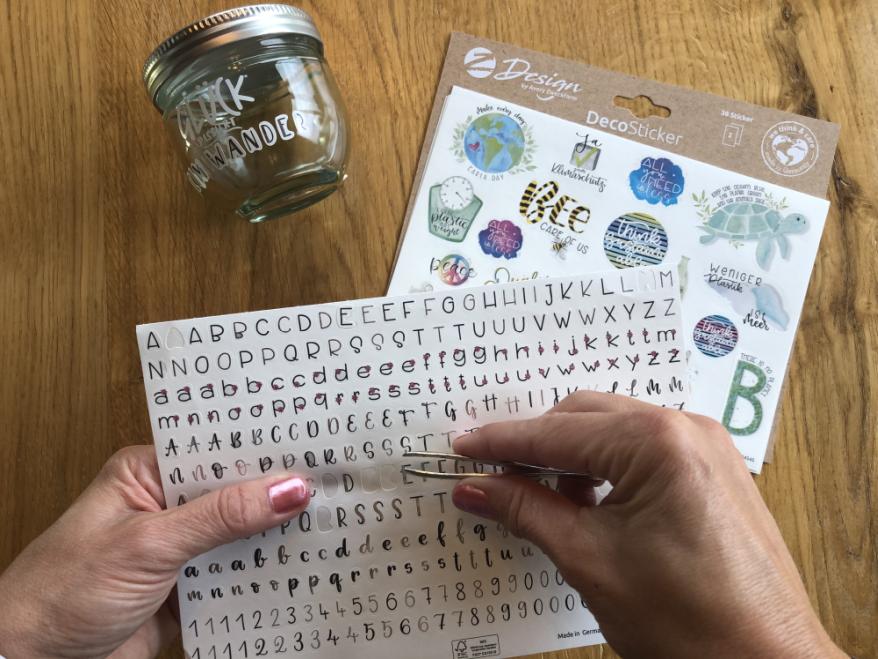 Eine Hand löst mit einer Pinzette Buchstabensticker von einem Bogen. außerdem sind ein Marmeladenglas und weitere Stickerbögen zu sehen.