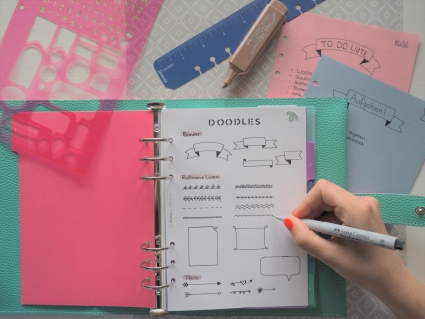 Doodles werden in einen Planner gemalt, rundherum Lineal, Schablone und einzelne Seiten.