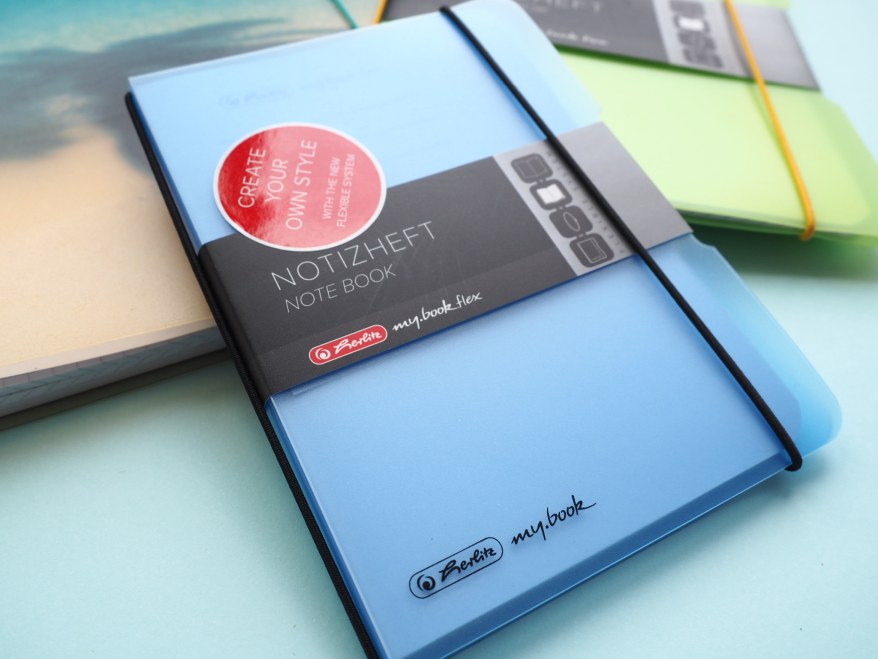 Blaues Herlitz mybook flex Notizheft, daneben ein grünes und ein beiges Heft.