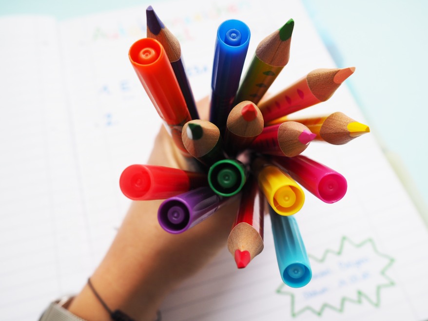 Hand hält bunte Stifte und Malstifte als Strauß über einem Notizheft.
