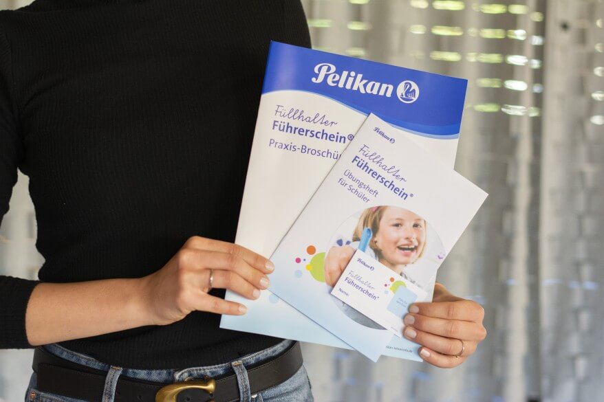 Eine Person hält diverse Unterlagen zum Thema Füllerführerschein der Marke Pelikan in der Hand: Eine Praxisbroschüre, ein Übungsheft für Schüler sowie den Füllhalter-Führerschein.