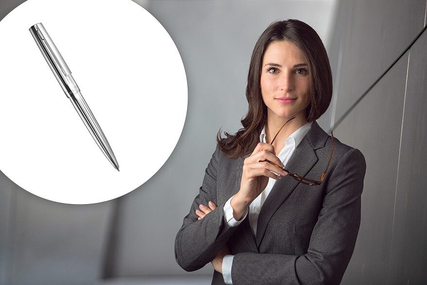 Eine Frau im Hosenanzug hält ihr Brille in der Hand und schaut in die Kamera. links neben ihr ist ein silberner Kugelschreiber design07 von Otto Hutt abgebildet.
