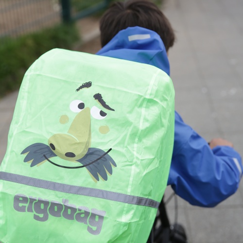 Ein Kind mit Regenjacke trägt einen Schulrucksack, der mit einer Regenhülle von ergobag geschützt ist. 