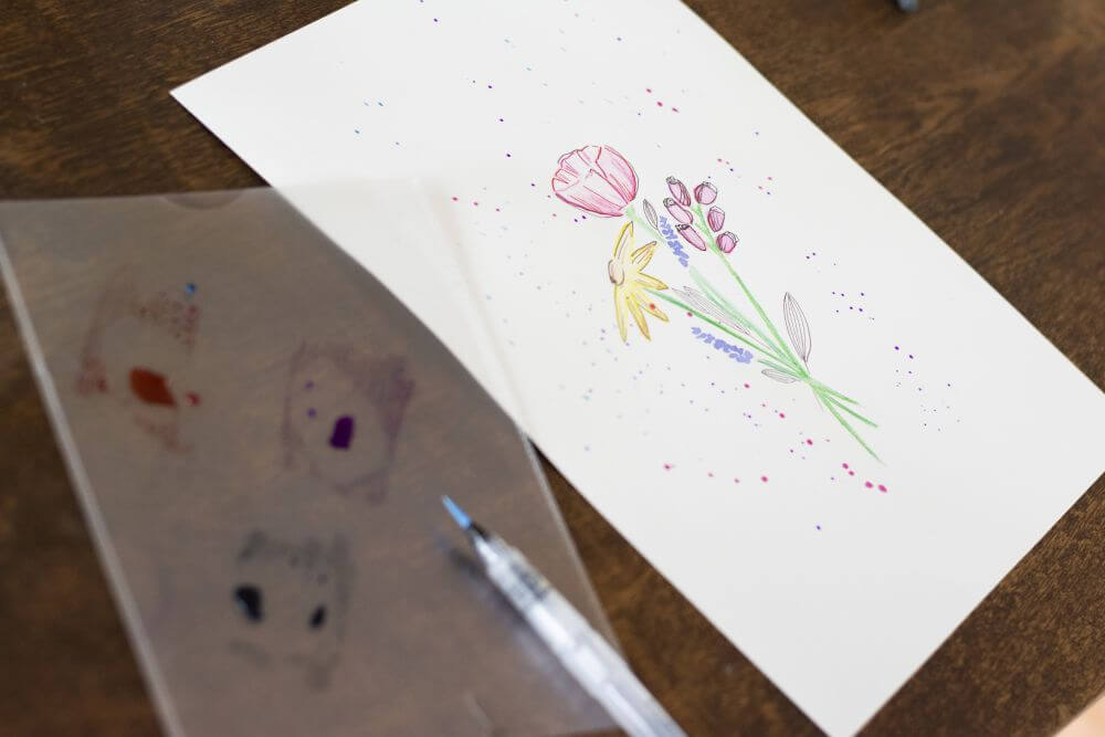 Auf einem Tisch liegt ein Blatt Papier mit gemaltem Blumenstrauß. Davor liegt eine Dokumentenhülle mit Farbflecken und Wassertankpinsel.