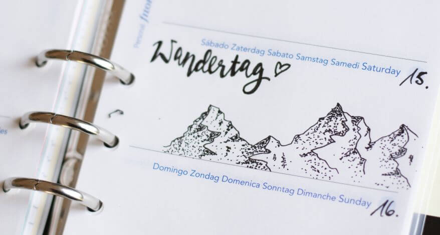 Nahaufnahme eines Kalenders von Filofax, in den mit Hilfe der Zeichentechnik Dotwork eine Bergkette gezeichnet wurde. Daneben steht als Lettering "Wandertag".