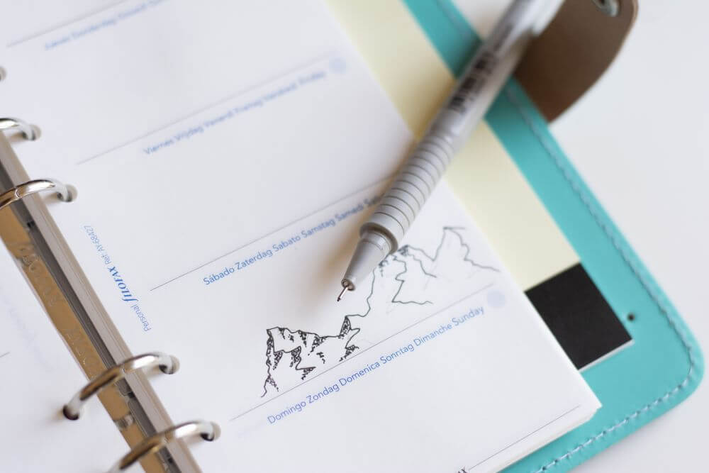 Ein aufgeschlagener Kalender von Filofax liegt auf einem Tisch, es entsteht gerade eine Bergkette, die mit der Zeichentechnik Dotwork mit einem Fineliner gezeichnet wird.