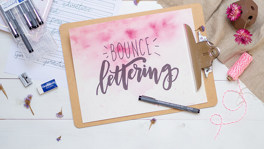Ein Klemmbrett mit dem Schriftzug Bounce Lettering auf rosa Aquarellgrund, liegt auf einem weißen Tisch. Drumherum liegen Pigment Liner und andere Schreibutensilien der Marke Staedtler. Foto: Johanna Rundel
