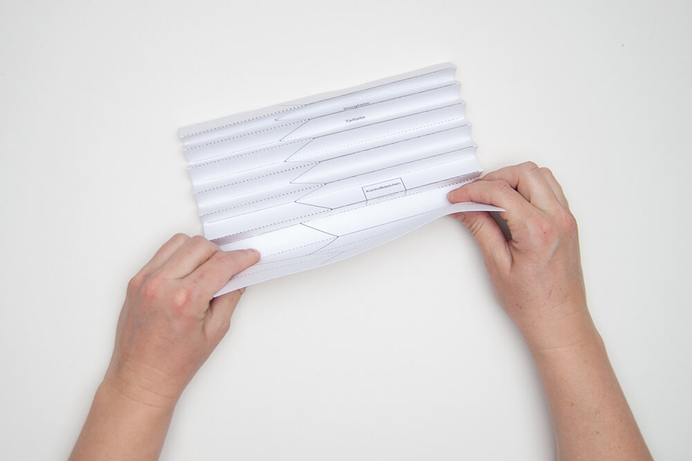 Pappvase: Hände falten die geraden Linien einer Faltvorlage auf einem Designpapier von Artoz zu einer „Ziehharmonika“.