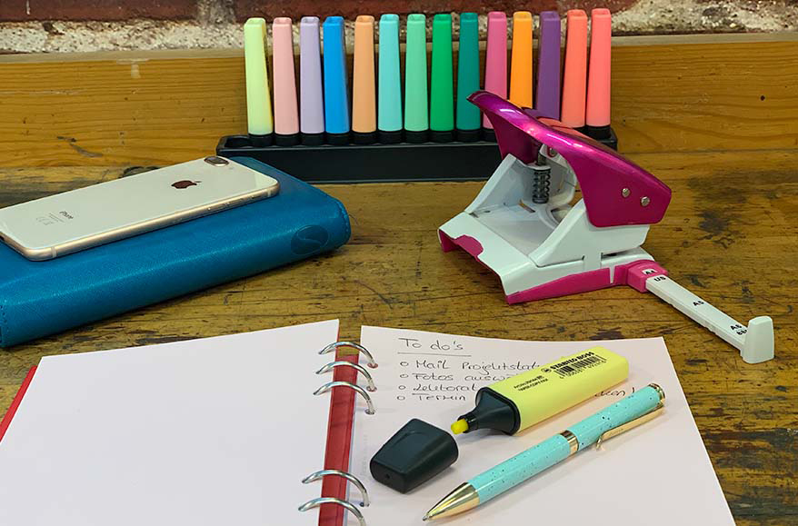 Man sieht einen bunt eingerichteten Arbeitsplatz. Im Hintergrund stehen zahlreiche Textmarker der Marke Stabilo, ein rosafarbenen Locher sowie ein Planer von Filofax auf dem ein Smartphone liegt.  Im Vordergrund liegt ein Notizbuch der Marke Filofax, in dem eine to-do-Liste geführt wird, sowie ein türkiser Kugelschreiber