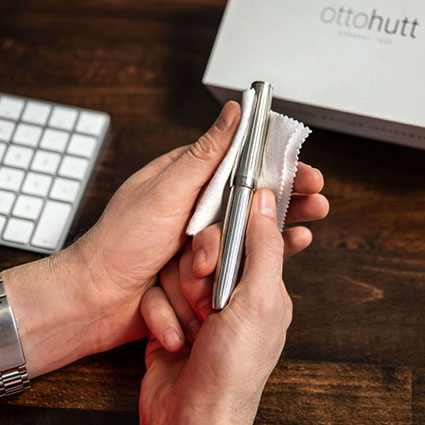 Ein Füller aus Silber der Marke Otto Hutt wird mit einem weichen Tuch gereinigt. Im Hintergrund liegen auf einem dunklen Holztisch eine Computertastatur und eine Geschenkverpackung von Otto Hutt.