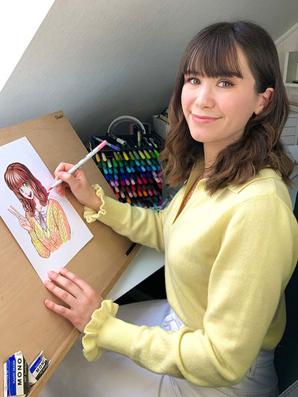 Ein Foto der Manga-Künstlerin Reyhan Yildirim. Sie sitzt vor ihrem Zeichenbrett und malt gerade ein Manga. Sie blickt lächelnd in die Kamera. Im Hintergrund sind ihre Zeichenmaterialen und Stifte von Tombow zu sehen. Foto: Reyhan Yildirim