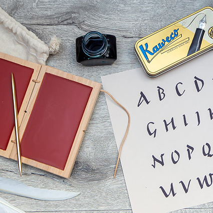 Auf einem rustikalen Holzuntergrund liegt ein Papier, auf dem mit römischer Schrift ein Alphabet geschrieben steht. Daneben liegt ein kleiner Jutebeutel, auf dem eine römische Wachstafel mit rotem Wachs und ein Griffel aus Bronze liegen. Oberhalb davon ist ein Tintenfass und eine Blechdose mit einem modernen Füllfederhalter von Kaweco zu sehen. Foto: Johanna Rundel