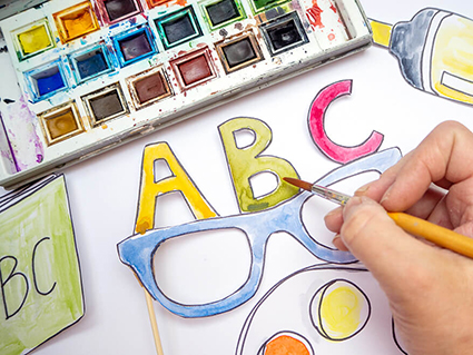 Jemand malt mit einem Pinsel und bunten Aquarellfarben ein Foto-Prop in Form einer Brille an. Auf der Brille sind die Buchstaben ABC zu sehen. Der verwendete Fotokarton ist von der Marke Heyda. Foto: Johanna Rundel