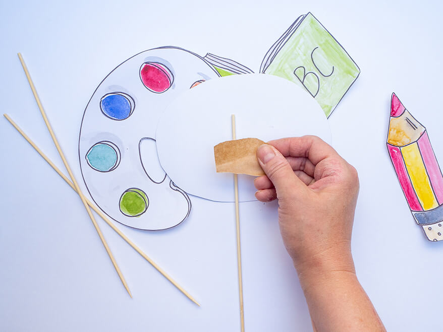 Jemand möchte Foto-Props für die Einschulung basteln und klebt dafür ein Holzstäbchen von Knorr-Prandell auf die Rückseite einer ausgeschnittenen Sprechblase aus Papier mit einem Klebeband fest. Foto: Johanna Rundel