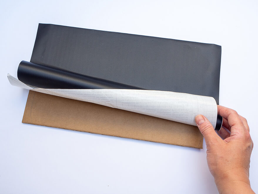 Jemand zieht schwarze Tafelfolie von Knorr-Prandell auf eine braune Pappe um daraus eine kleine Schultafel als Foto-Prop für die Einschulung zu basteln. Foto: Johanna Rundel