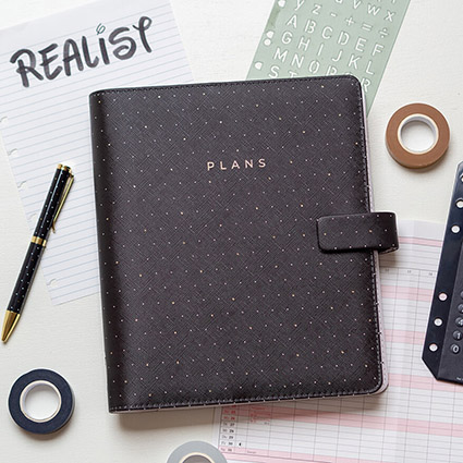Auf einem weißen Untergrund liegt ein schwarzer Lederplaner mit goldenen Pünktchen von Filofax. Darauf steht in goldenen Buchstaben "Plans" steht. Darunter liegt ein Zettel mit der Aufschrift "Realist". Drumherum sind verschiedene Materialien verteilt: Kalenderblätter, Stift, Taschenrechner, Washitapes in neutralen Farben und eine Schablone. Foto: Johanna Rundel