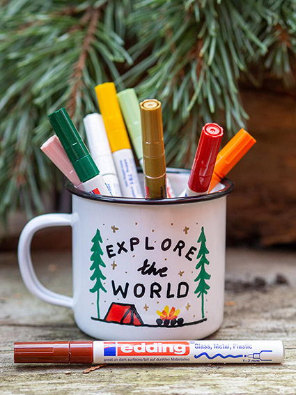 Auf einem rustikalen Holzuntergrund steht eine weiße Emailletasse mit schwarzem Rand. Sie wurde mit Glanzlackmarkern von edding mit einem Campingmotiv bemalt. Darauf zu sehen sind zwei grüne Tannen, ein rotes Zelt, ein Lagerfeuer und der Spruch "Explore the World". In und vor der Tasse befinden sich die zum Malen verwendeten Stifte. Im Hintergrund ist ein Kieferzweig zu sehen. Foto: Johanna Rundel
