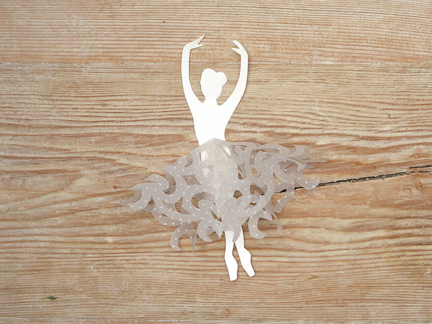 Aus einem hellen Papier von Artoz wurde die Silhouette einer Tänzerin ausgeschnitten. Aus Transparentpapier von Artoz wurde in Scherenschnitttechnik eine Schneeflocke ausgeschnitten und als Tutu um die Taille der Ballerina gelegt. Der Untergrund ist ein rustikales, helles Holzbrett. Foto: Johanna Rundel
