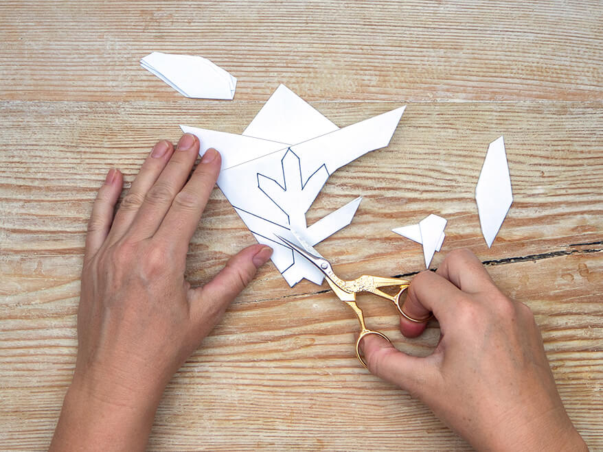 Jemand schneidet mit einer kleinen, goldenen Schere den Umriss einer einfachen Schneeflocke aus einem zu einem Dreieck gefalteten Papier von Artoz aus. Die Papierschnipsel liegen daneben. Der Untergrund ist ein rustikales, helles Holzbrett. Foto: Johanna Rundel