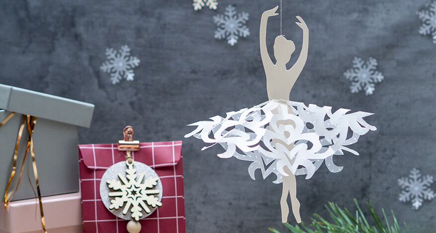 Vor einem dunklen Hintergrund hängt eine Ballerina aus weißem Papier von Artoz. Das Tutu ist aus Transparentpapier mit einer Scherenschnitttechnik zur Schneeflocke gearbeitet. Im Hintergrund hängen weitere Papierschneeflocken. Einige Tannenzweige dienen als zusätzliche Deko. Foto: Johanna Rundel