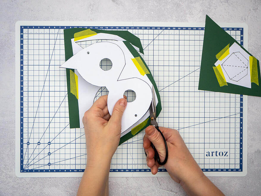 Jemand schneidet mit einer schwarzen Schere entlang einer aufgeklebten Vorlage eine Vogelmaske aus einem dunkelgrünen Fotokarton. Der Untergrund ist eine helle, faltbare Schneidematte mit Linien von Artoz. Foto: Johanna Rundel