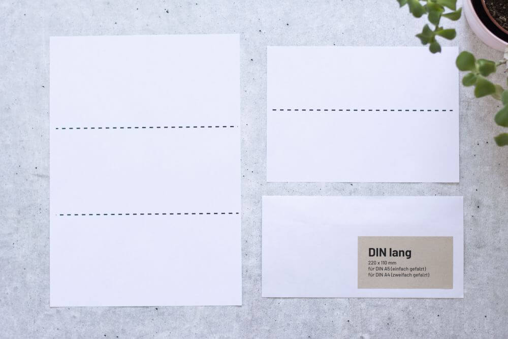 Auf grauem Grund liegt ein DIN A4-Papier mit zwei gestrichelten Markierungen, die das Papier in Drittel teilt und ein DIN A5-Papier mit einer gestrichelten Markierung in der Mitte, dazu ein passender Briefumschlag DIN lang. Auf dem Briefumschlag ist ein Aufkleber aufgebracht mit folgenden Infos: DIN lang, 220x110 Millimeter, für DIN A5 (einfach gefalzt), für DIN A4 (zweifach gefalzt). Rechts oben ragt der Teil einer Pflanze ins Bild.