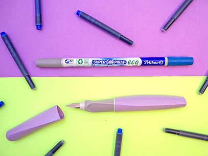 Das Bild ist zweigeteilt. Die obere Hälfte hat einen lavendelfarbenen Untergrund. Die untere Hälfte ist hellgelb. Auf der lila Seite liegt ein Tintenlöscher von Pelikan, auf der gelben ein lavendelfarbener Füller. Drumherum liegen in unregelmäßigen Abständen lange, blaue Tintenpatronen. Foto: Johanna Rundel