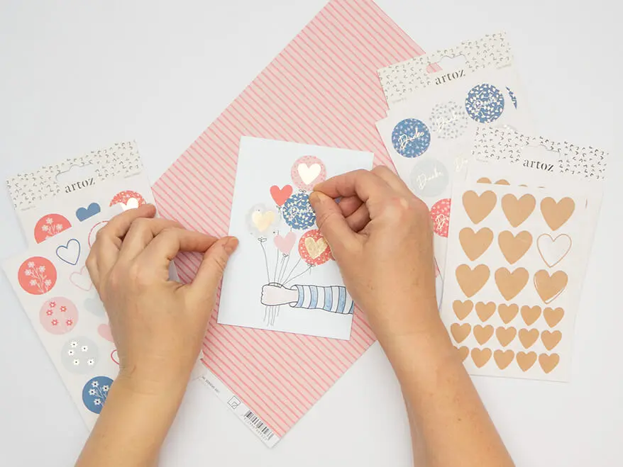 Jemand klebt Aufkleber als "Luftballonwolke" über einen ausgestreckten, gezeichneten Arm auf einer weißen Klappkarte von Artoz. Weitere Aufkleberbögen liegen um die Karte verteilt. Der Untergrund ist weiß. Foto: Johanna Rundel