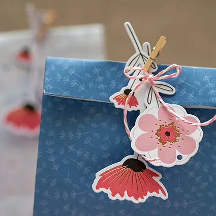 Nahaufnahme einer blau-geblümten Geschenktüte von Artoz. Es ist die Verschluss-Faltung zu sehen, die mit einer kleinen Holzwäscheklammer fixiert wurde. In der Klammer stecken zwei Geschenkanhänger in Blütenform, die mit etwas rosa-weiß-gestreiftem Bäckergarn zusammengehalten werden. Foto: Johanna Rundel