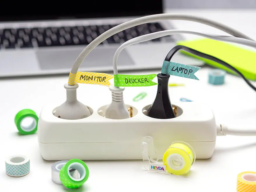 Vor einem grauen Laptop auf einem weißen Schreibtisch steht eine weiße Mehrfachsteckdose. Die drei Stecker, die darin stecken, wurden mit Fähnchen aus buntem Washitape markiert. Es stehen die Worte Monitor, Drucker und Laptop darauf. Im Vordergrund sind mehrere kleine Rollen farbiges Washitape von Heyda zu sehen. Foto: Johanna Rundel