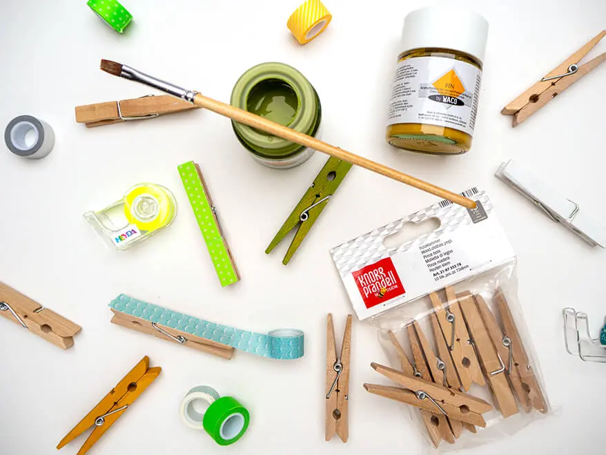 Auf einem weißen Untergrund liegen verschiedene Bastelmaterialien: Wäscheklammern von Knorr Prandell, Washitape von Heyda, Acrylfarbe von Waco sowie ein Pinsel. Foto: Johanna Rundel