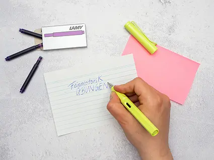 Jemand schreibt mit einem hellgrünen Füller von Lamy „Feinmotorik Übungen“ auf eine große Karteikarte. Darüber ist ein Päckchen mit lila Tintenpatronen zu sehen.