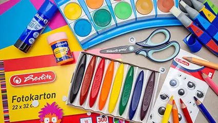 Auf einem grauen Untergrund sind verschiedene Bastelmaterialien und Schulbedarf der Marken Herlitz und Pelikan verteilt. Foto: Johanna Rundel