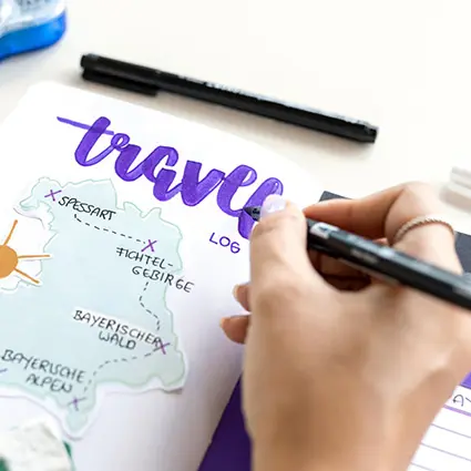 Jemand schreibt mit einem lila Brush Pen von Tombow das Wort "travelling" über eine Landkarte in einem Bullet Journal.