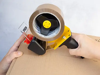 Jemand klebt ein Paket mit braunem Versand-Klebeband aus einem großen Klebebandabroller zu. Foto: Johanna Rundel