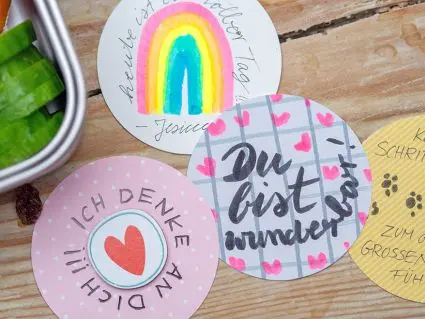 Bunte, runde Affirmationskarten mit motivierenden Sprüchen und Bildern liegen neben einer offenen Edelstahlbrotdose von ergobag. Foto: Johanna Rundel