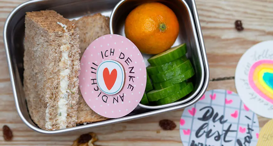 Eine gefüllte Edelstahlbrotdose von ergobag auf einem Holzbrett. In und um die Dose herum liegen verschiedene, runde Affirmationskarten mit motivierenden Sprüchen und Bildern. Foto: Johanna Rundel 