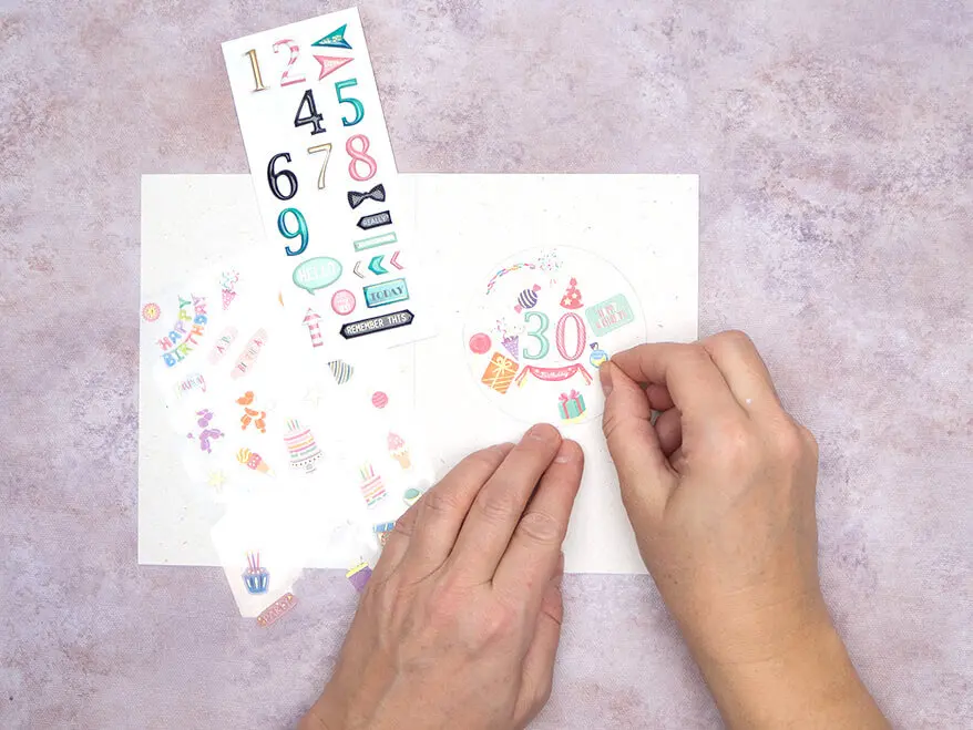 Jemand klebt Zahlen- und Mini-Geburtstagsaufkleber von Rössler auf eine Blanko-Klappkarte. Foto: Johanna Rundel 