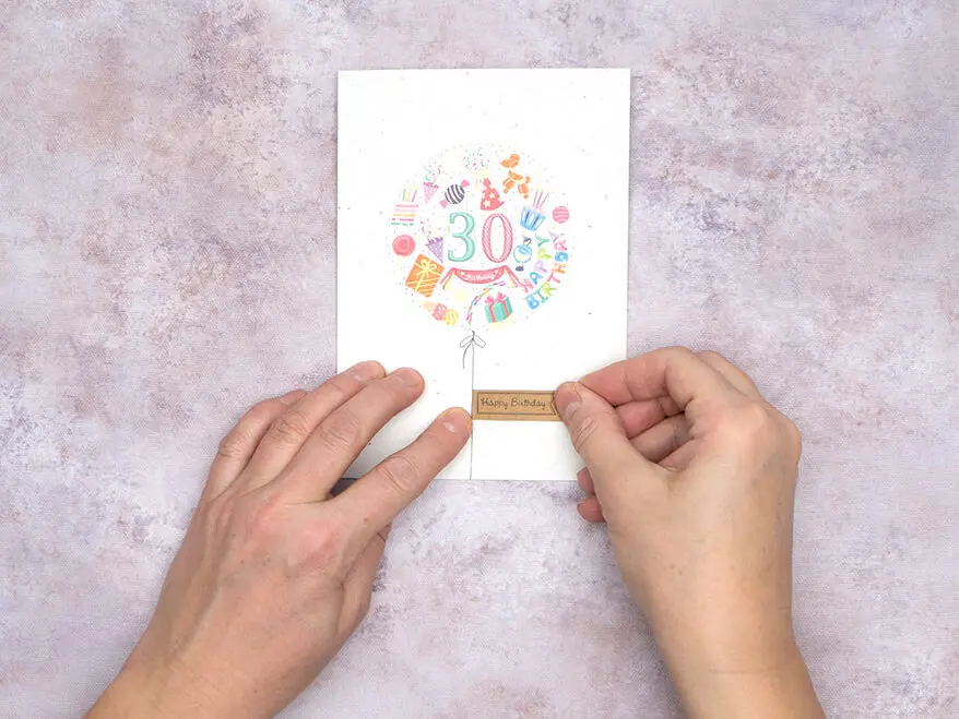 Jemand klebt einen „Happy Birthday“-Aufkleber aus Naturkork von Rössler auf eine gestaltete Glückwunschkarte zum 30. Geburtstag. 