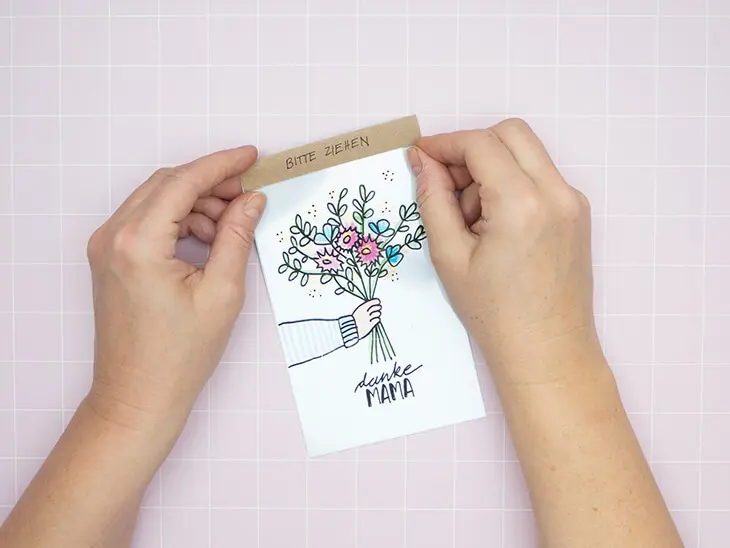 Jemand klebt ein Kraftpapier-Streifen mit der Aufschrift "bitte ziehen" an die Oberseite einer Schiebekarte zum Muttertag. Foto: Johanna Rundel