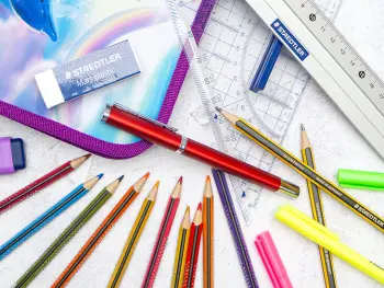 Verschiedene Schulmaterialien, wie Stifte, Textmarker, Lineal und Radiergummi von STAEDTLER liegen verteilt auf einem hellgrauen Untergrund. Foto: Johanna Rundel