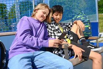 Zwei Jugendliche sitzen gechillt auf einer Bank vor einem Sportcourt und schauen gemeinsam auf ein Handy.