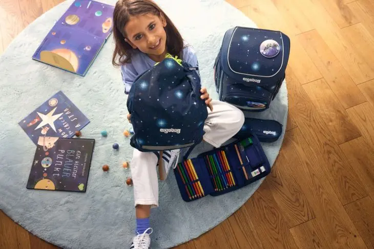 Ein Mädchen sitzt auf dem Boden neben einem Schulranzen von ergobag, umgeben von Schulzubehör