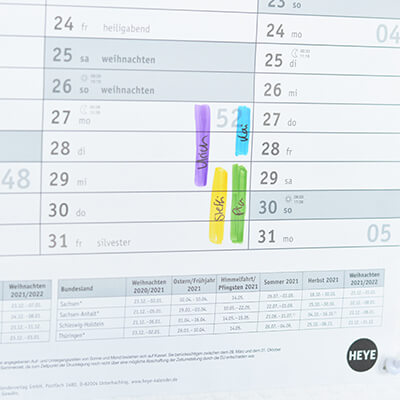 Detailaufnahme eines Posterplaners: Man sieht den Ausschnitt eines Wandkalenders, in dem mit farbigen Balken die Namen der Mitarbeiter eingetragen sind, die an den jeweiligen Tagen Urlaub haben. 
