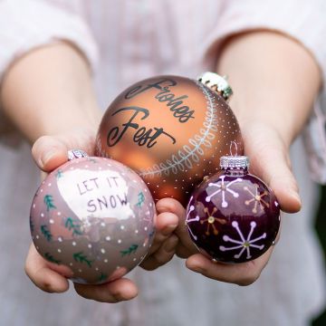 Eine Frau hält drei beletterte Weihnachtskugeln in dunklem Beerenton, Burgunderrot und Pflaumenlila ind die Kamera..